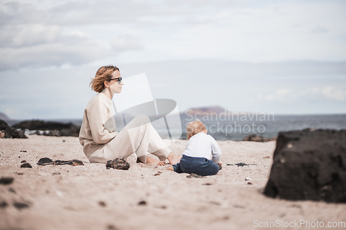 Image of Mother enjoying winter beach vacations playing with his infant baby boy son on wild volcanic sandy beach on Lanzarote island, Canary Islands, Spain. Family travel and vacations concept