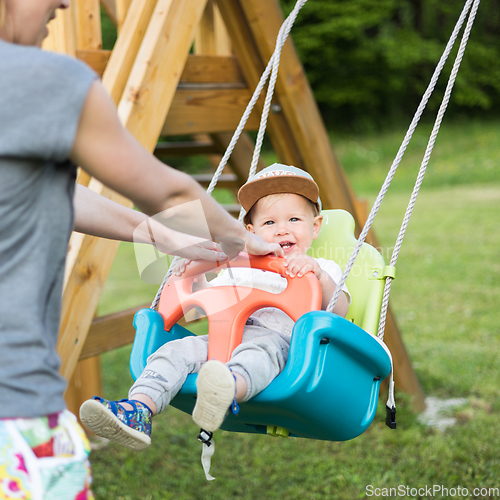 Image of Mother pushing her infant baby boy child on a swing on playground outdoors.