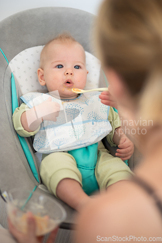 Image of Mother spoon feeding her baby boy infant child in baby chair with fruit puree. Baby solid food introduction concept.