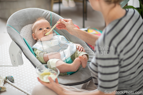 Image of Mother spoon feeding her baby boy infant child in baby chair with fruit puree. Baby solid food introduction concept.