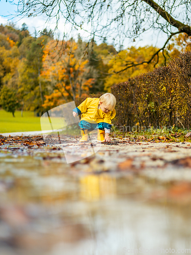 Image of Sun always shines after the rain. Small bond infant boy wearing yellow rubber boots and yellow waterproof raincoat walking in puddles in city park on sunny rainy day.