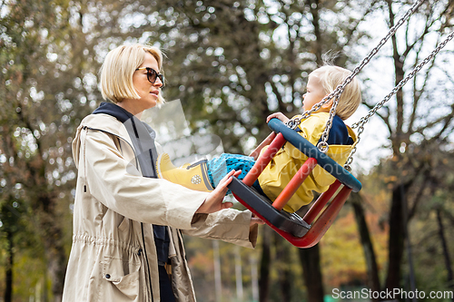 Image of Mother pushing her infant baby boy child wearing yellow rain boots and cape on swing on playground outdoors on cold rainy overcast autumn day in Ljubljana, Slovenia