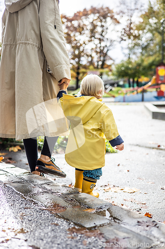 Image of Small bond infant boy wearing yellow rubber boots and yellow waterproof raincoat walking in puddles on a overcast rainy day holding her mother's hand. Mom with small child in rain outdoors.