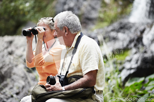 Image of Senior, couple and binocular in forest for tourist, sightseeing or vacation with waterfall and scenery. Elderly, man and woman in woods or nature for holiday, adventure or experience with backpack