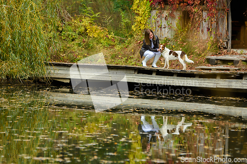 Image of Young beautiful girl petting a dog near a lake in an autumn park