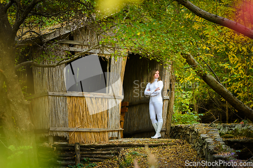 Image of A young beautiful girl in white clothes stands near an old abandoned house in the forest