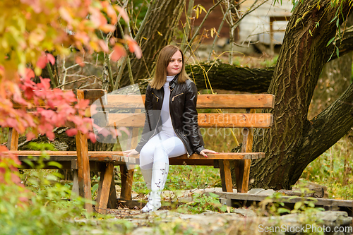 Image of A young beautiful girl sits on a bench in an autumn park and looks at the surrounding nature