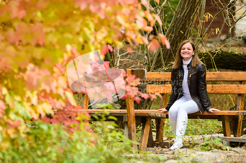 Image of A young beautiful girl sits on a bench in an autumn park and looks happily into the frame