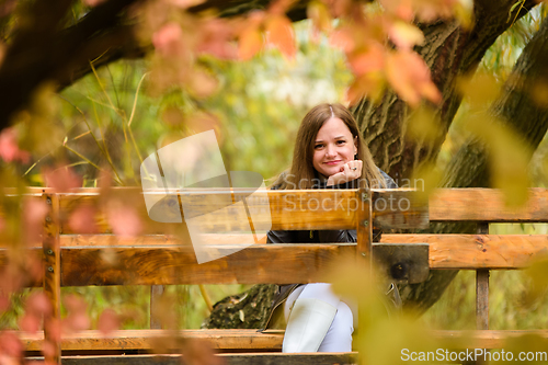 Image of A young beautiful girl sits on a bench in an autumn park and looks into the frame, smiling.