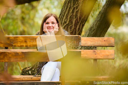 Image of A young beautiful girl sits on a bench in an autumn park and looks cheerfully into the frame