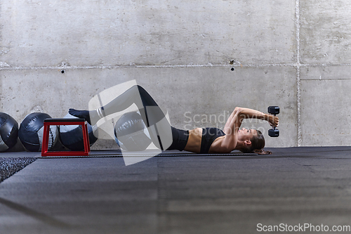 Image of A fit woman is lying on the gym floor, performing arm exercises with dumbbells and showcasing her dedication and strength.