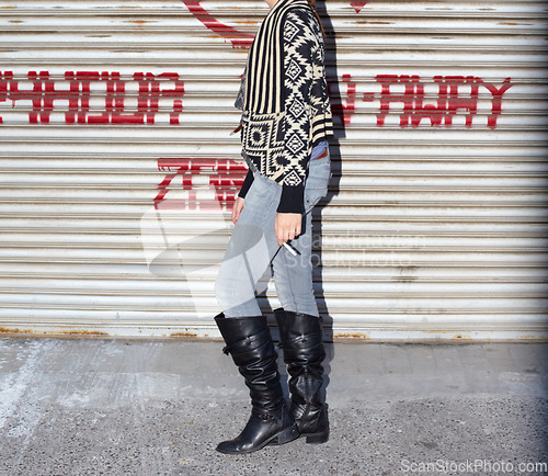 Image of Boots, fashion and body of young woman by garage door for stylish, cool and edgy outfit with jeans. Legs, feet and zoom of female person with cigarette and trendy grungy style by steel background.