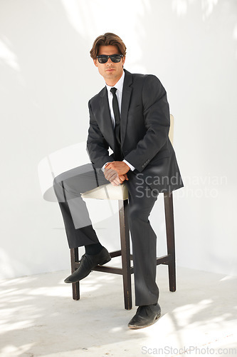 Image of Portrait, chair and sunglasses with a business man outdoor on a white background for corporate style. Work, professional and formal suit with a young employee sitting on a stool for trendy fashion