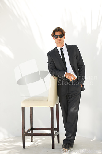 Image of Portrait, fashion and a business man with a chair in studio on a white background for corporate style. Handsome, sunglasses and formal with a confident young professional employee leaning on a seat