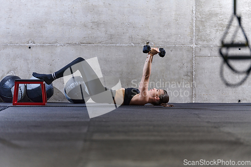 Image of A fit woman is lying on the gym floor, performing arm exercises with dumbbells and showcasing her dedication and strength.