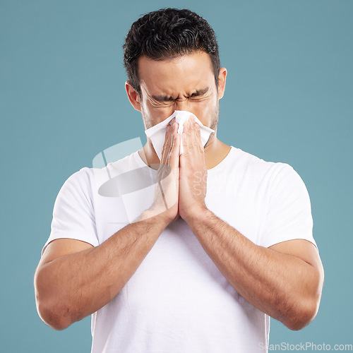 Image of Handsome young mixed race man blowing his nose while standing in studio isolated against a blue background. Hispanic male suffering from cold, flu, sinus, hayfever or corona and using a facial tissue