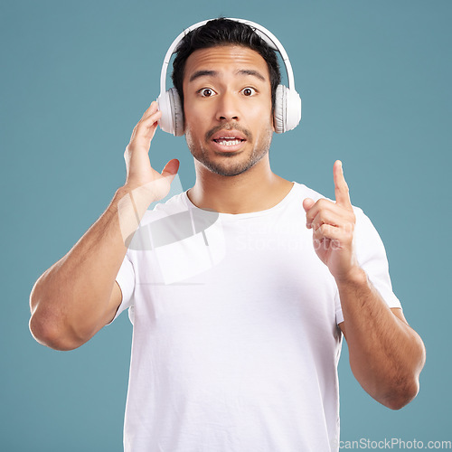 Image of Handsome young mixed race man looking surprised and listening to music while standing in studio isolated against a blue background. Hispanic male remembering a song while using wireless headphones