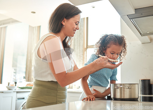 Image of Mother giving little daughter a wooden spoon to taste. Mother and daughter cooking together in the kitchen. Child tasting flavour in food