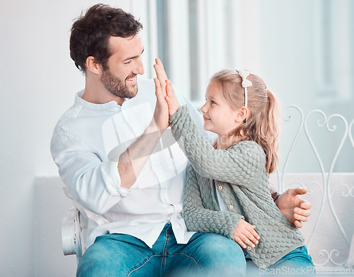 Image of Cheerful caucasian young father giving his little daughter a high five to support and motivate her while sitting on a chair together at home. Man joining hands with his child enjoying a day together