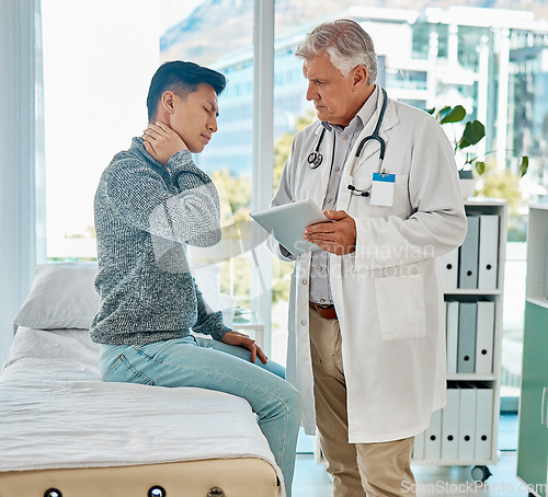 Image of Mature medical doctor standing and using a digital tablet while checking on his injured asian patient in a clinic. Young mixed race man suffering from a painful neck in a hospital. Tension and sprain