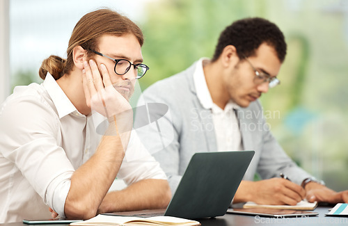 Image of When will this day end. a young businessman looking bored while working on a laptop in an office with his colleague in the background.