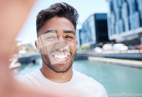 Image of Young mixed race sportsman smiling and holding mobile phone with hand to take a selfie while out for a run or jog along a water canal in the city. Happy male athlete taking self-portrait during worko