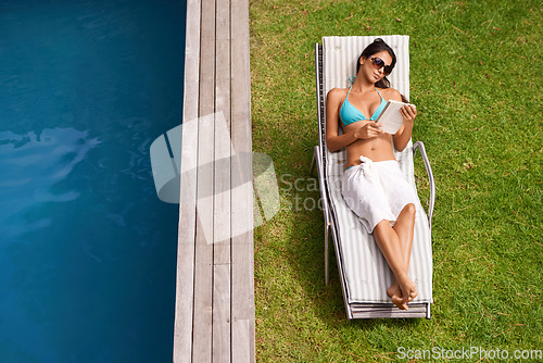 Image of Reading while soaking up some sun. A beautiful young woman reading a book while relaxing beside a swimming pool.