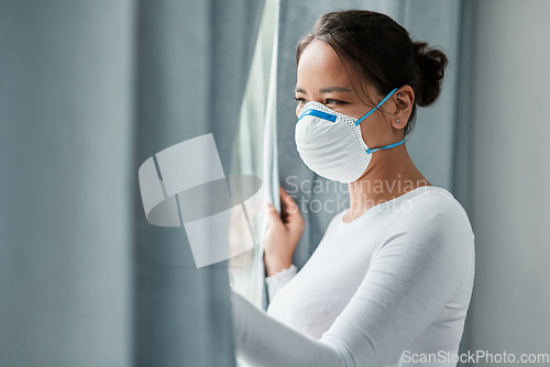 Image of The lockdown has affected our lives in so many ways. a young woman wearing a mask while looking out a window at home.