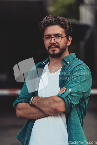 Image of A successful young businessman in a shirt, with crossed arms, poses outdoors, confident expression on his face.