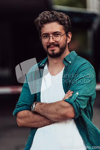 Image of A successful young businessman in a shirt, with crossed arms, poses outdoors, confident expression on his face.