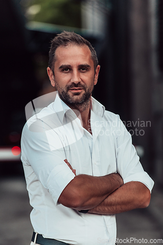 Image of A successful businessman in a white shirt, with crossed arms, poses outdoors, confident expression on his face.