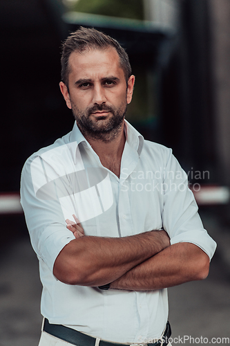 Image of A successful businessman in a white shirt, with crossed arms, poses outdoors, confident expression on his face.