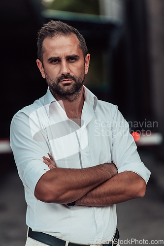 Image of A successful businessman in a white shirt, with crossed arms, poses outdoors, confident expression on his face.