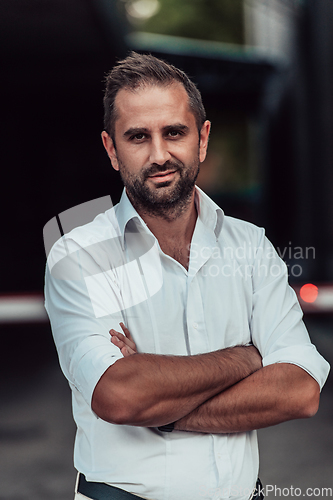 Image of A successful businessman in a white shirt, with crossed arms, poses outdoors, confident expression on his face.