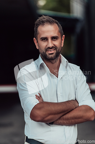 Image of A successful businessman in a white shirt, with crossed arms, poses outdoors, confident expression on his face.