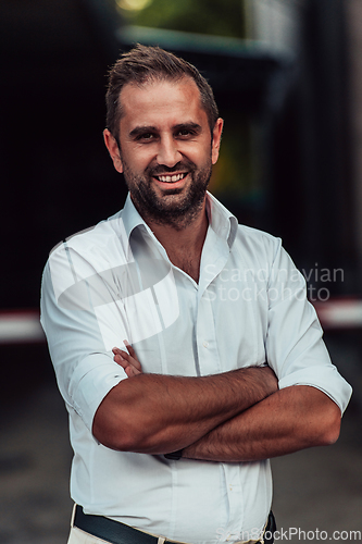 Image of A successful businessman in a white shirt, with crossed arms, poses outdoors, confident expression on his face.