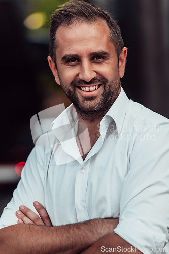 Image of A successful businessman in a white shirt, with crossed arms, poses outdoors, confident expression on his face.