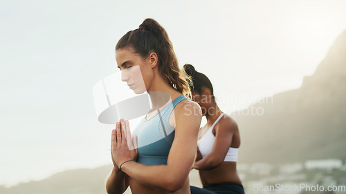 Image of Meditation is being in tune with our inner energy source. two attractive young women sitting and meditating together on the beach during an overcast day.