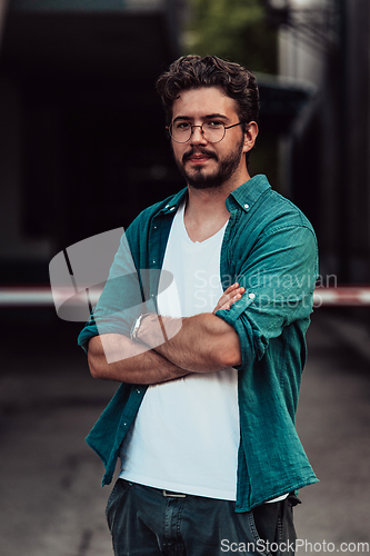 Image of A successful young businessman in a shirt, with crossed arms, poses outdoors, confident expression on his face.