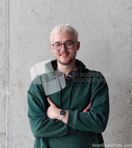 Image of A man with blue hair, eyeglasses, and a green sweatshirt confidently poses with his arms crossed against a gray background, showcasing his fashionable and unique style.