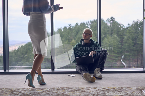 Image of A young blond man in a modern office is sitting by the window, engrossed in his work on a laptop while talking to a female colleague