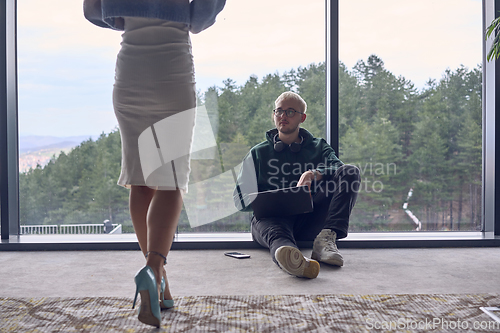 Image of A young blond man in a modern office is sitting by the window, engrossed in his work on a laptop while talking to a female colleague