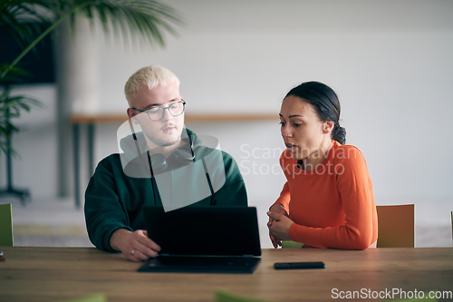 Image of A young entrepreneurial couple sits together in a large, modern office, engaged in analyzing statistics and data on their laptop