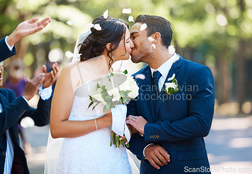 Image of Happy bride and groom sharing a kiss while surrounded by guests and being showered with confetti rose petals after wedding ceremony