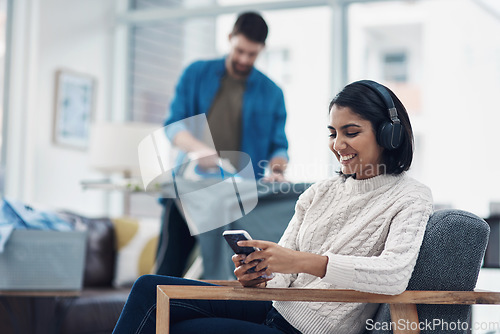 Image of Today it’s hubby’s turn, tomorrow it’s mine. a young woman using a smartphone and headphones while her husband irons clothing in the background.