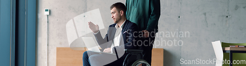 Image of Wide crop of businessman in a wheelchair in a fashionable office using a laptop while behind him is his business colleague who gives him support