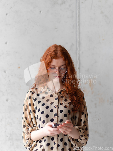 Image of A young entrepreneur with captivating orange hair is striking a confident pose while holding a smartphone in front of a stylish gray wall.