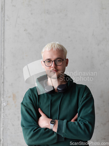 Image of A man with blue hair, eyeglasses, and a green sweatshirt confidently poses with his arms crossed against a gray background, showcasing his fashionable and unique style.