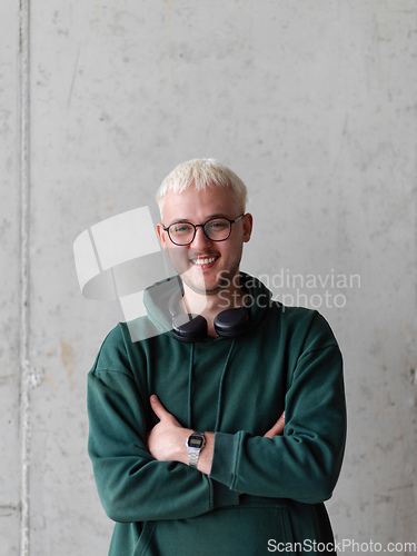 Image of A man with blue hair, eyeglasses, and a green sweatshirt confidently poses with his arms crossed against a gray background, showcasing his fashionable and unique style.