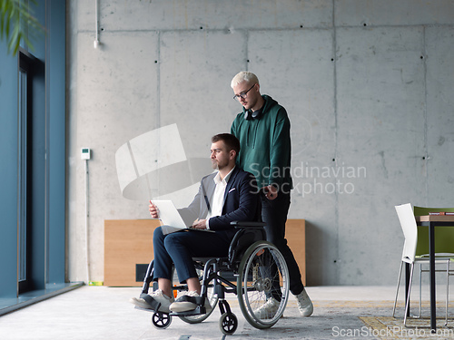 Image of A businessman in a wheelchair in a fashionable office using a laptop while behind him is his business colleague who gives him support